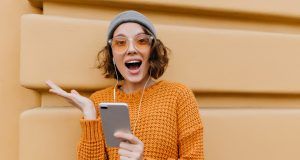 Active girl in trendy cozy attire posing with happy face expression, holding smartphone. Excited young lady in knitted outfit waving hand in front of building after phone call..