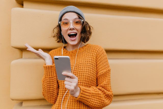 Active girl in trendy cozy attire posing with happy face expression, holding smartphone. Excited young lady in knitted outfit waving hand in front of building after phone call..