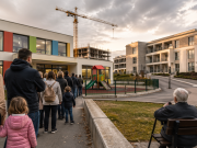 Roditelji s djecom i starije osobe stoje u redu ispred novog vrtića dok se u pozadini vidi moderna zgrada i gradilište, ilustrirajući nedostatak mjesta unatoč ulaganjima