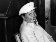 Starija crno-bijela fotografija Tita koji sjedi naslonjen na stolac, nasmiješen s cigarom u ustima, u interijeru s drvenim zidom u pozadini.