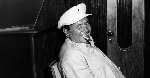 Starija crno-bijela fotografija Tita koji sjedi naslonjen na stolac, nasmiješen s cigarom u ustima, u interijeru s drvenim zidom u pozadini.